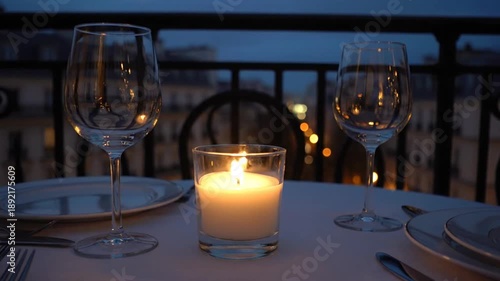 Romantic dinner setup with wine glasses and lit candle on a balcony overlooking city lights at dusk.