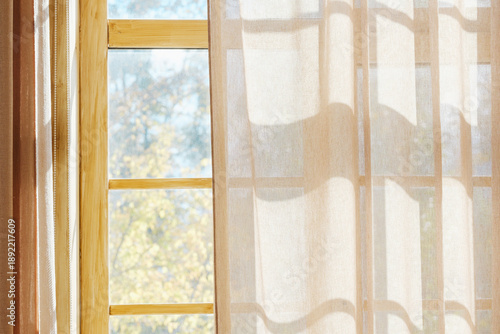 Sunlight through sheer curtains illuminates a wooden window frame with autumn trees outside