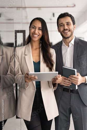 Happy professional multinational teammates posing in workspace. Two project managers working together using tablet, holding personal organizer, standing in office, looking at camera. Teamwork, unity