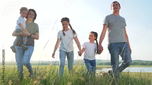 Family walk in meadow by lake with father and mother holding daughter and son while parent carries toddler child and grass sways under sky and distant hill reflects on calm water joyful outing
