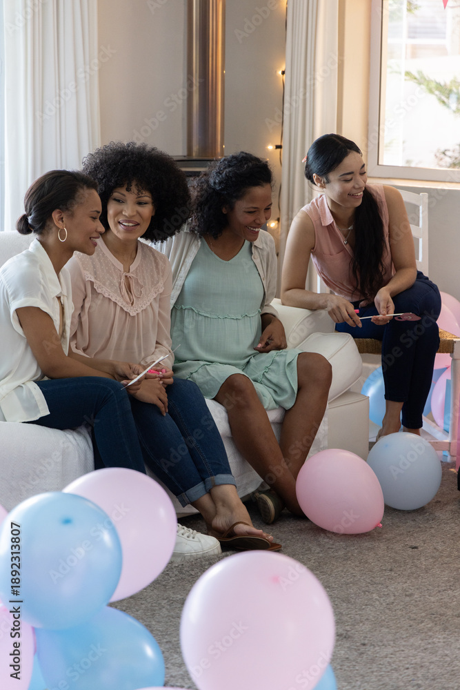 Fototapeta premium Diverse female friends chatting and examining cards on sofa at home, balloons, string lights