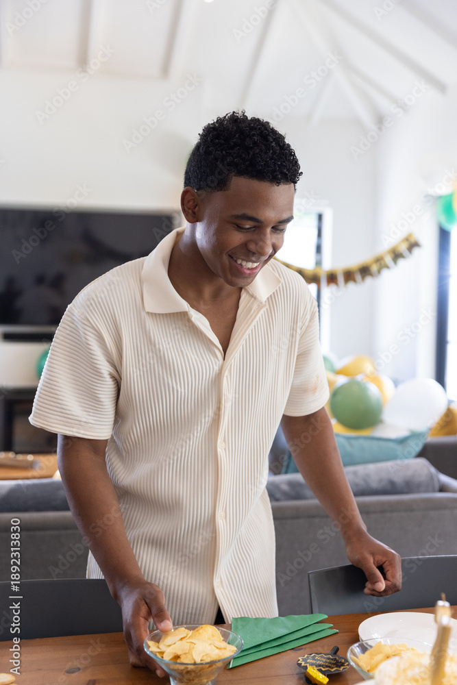 Fototapeta premium African american male leaning on table at home placing bowl of tortilla chips near green napkins