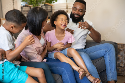 Diverse family sitting on gray sofa in living room, father tickling daughter and mother watching