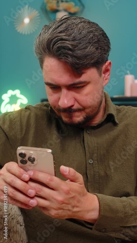 Middle-aged man at home types smartphone message pauses erases rewrites showing hesitation and thought about words. Young adult guy on sofa holds mobile phone pondering decision seeking best wording