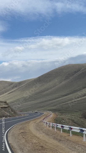 Scenic winding mountain road with cars under a cloudy sky, showcasing vast, rolling hills and distant horizons. A journey through a tranquil, expansive landscape.