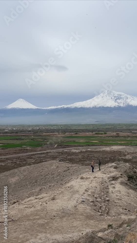 Mount Ararat view from a rocky ridge with two individuals overlooking distant green fields under a cloudy sky, showcasing a vast natural landscape.Armenia, Yerevan 5.02.2023