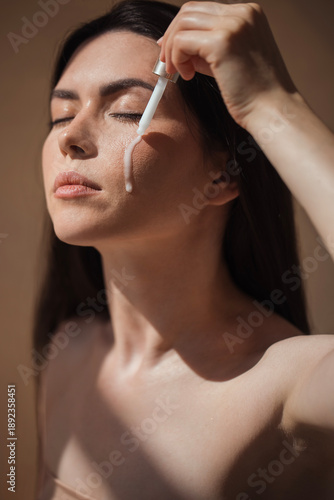 Young woman applying serum to her face with a dropper, showcasing a skincare routine, illuminated by soft natural light, emphasizing beauty and self-care practices