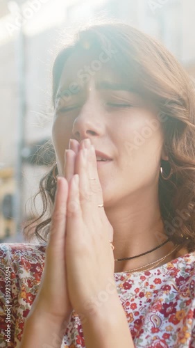 Close-up portrait of serious sad woman praying with closed eyes to God asking for blessing, hope help, forgiveness outdoors. Girl clasping hands wishing luck in urban city street. Religion lifestyles