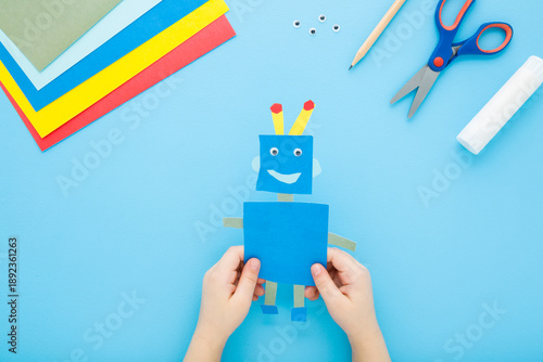 Little child boy hands holding and showing robot shape from colorful application paper on light blue table background. Pastel color. Paper craft. Closeup. Point of view shot. Flat lay. Top down view.
