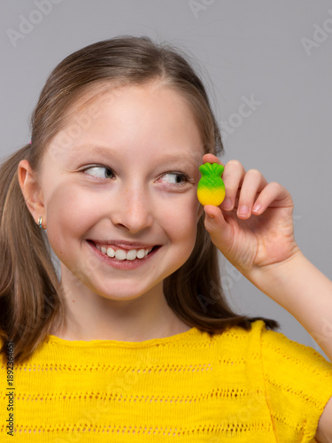 smiling girl in a close-up shot against a light neutral background. She has fair skin and light brown hair styled in two low pigtails. She is wearing a bright yellow knitted T-shirt that matches well 