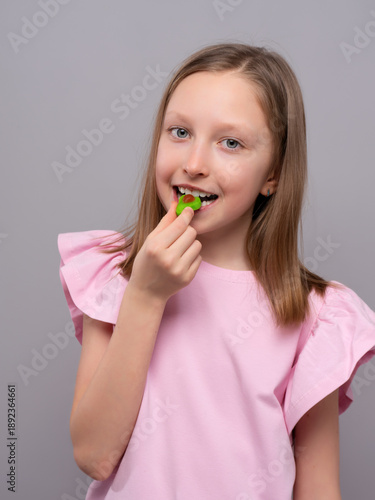 smiling young girl with fair skin and straight light brown hair is enjoying a gummy candy against a light gray studio background. She is wearing a soft pink shirt with ruffled sleeves and looking at t