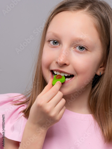 smiling young girl with fair skin and straight light brown hair is enjoying a gummy candy against a light gray studio background. She is wearing a soft pink shirt with ruffled sleeves and looking at t