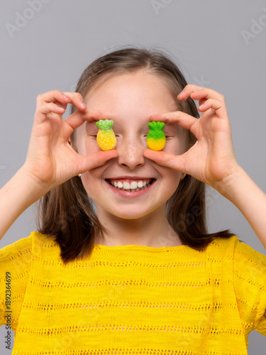 A cheerful young girl with light skin and brown hair in pigtails is posing against a light gray studio background. She is wearing a bright yellow knitted shirt and smiling broadly. The child playfully