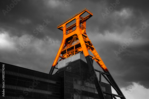 Steel framework of a historic coal mine winding tower in Essen (Ruhr region, Germany) in the evening sun. Bright orange structure isolated against a black-and-white background with dramatic cloudy sky
