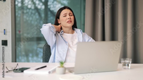 Woman experiences sudden, intense neck pain working on laptop, expressing discomfort and frustration. Seeks relief by massaging sore neck muscles, clear signs of strain from prolonged computer use.