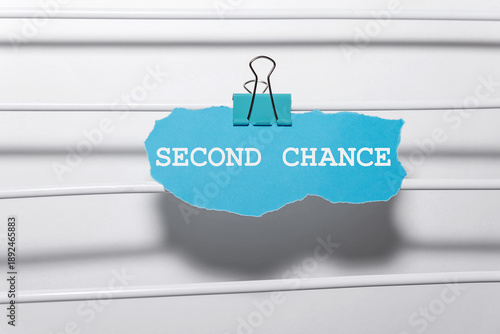 A blue paper with the words 'SECOND CHANCE' clipped to a white surface. The background is simple and minimalistic, emphasizing the message.