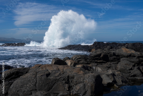 A large wave crashes forcefully against the shore of volcanic rocks