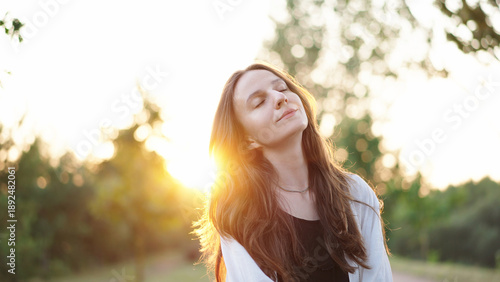 Portrait calm, beautiful young woman with closed eyes smiling at evening sunset, relieving stress after hard day in park. Serene brunette woman relaxes in the fresh air, enjoying the peace and quiet.
