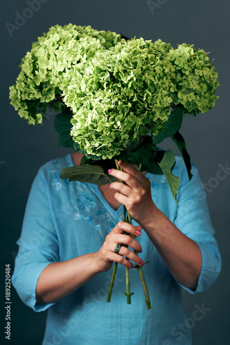 Woman holding green hydrangea bouquet covering face against dark studio background portrait