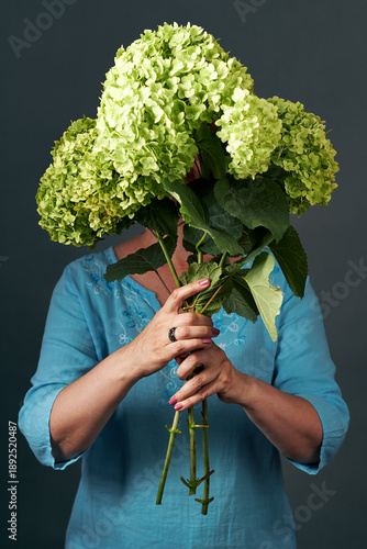 Woman holding green hydrangea bouquet covering face against dark studio background portrait