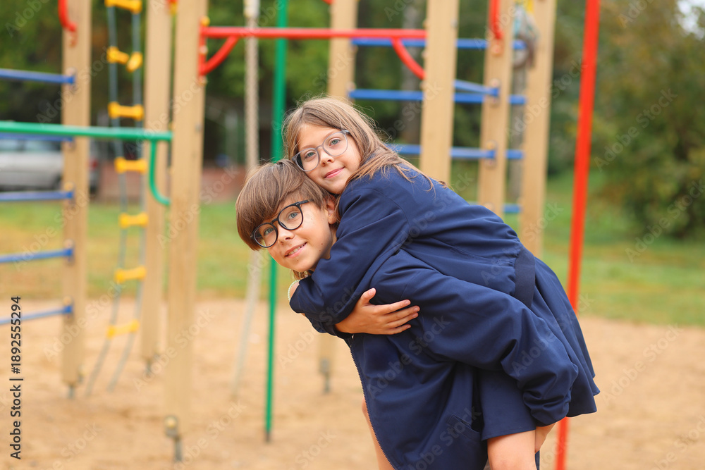 Obraz premium A boy gives a girl a piggyback ride on the playground, both smiling in school uniforms, radiating joy, trust, and playful friendship during recess.