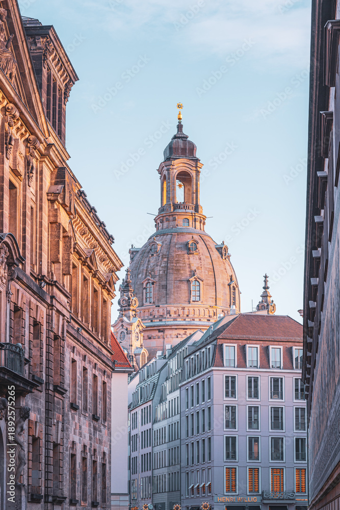 Fototapeta premium Frauenkirche stands tall in Dresden. The view captures the church's dome rising above nearby buildings. It is early morning, and the sky is clear. Visitors explore the area and take photos.