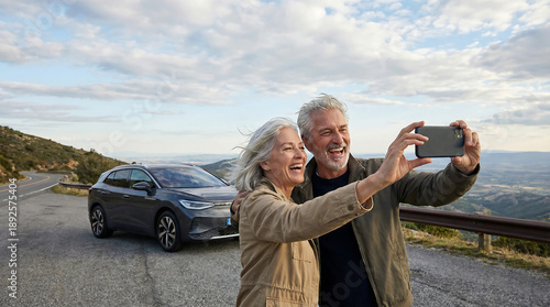 Happy senior couple taking smartphone selfie on scenic mountain overlook with electric car road trip travel adventure freedom and active retirement lifestyle