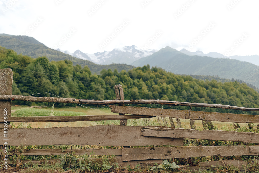 Fototapeta premium Fence meadow mountains forest rural landscape countryside rustic nature wooden A rustic wooden fence in green meadow with forest and distant snow mountains under cloudy sky