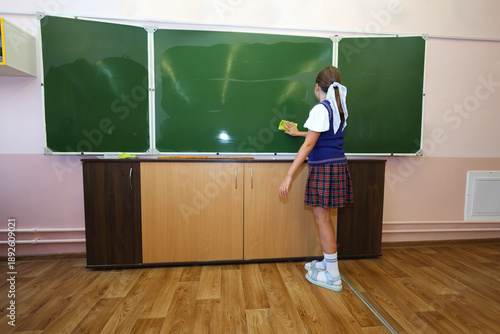 A girl in school uniform wipes the chalkboard with a green cloth, responsible, diligent, and contributing to classroom upkeep during or after lessons.