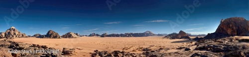 A panoramic desert landscape depicts a vast, sandy plain under a clear blue sky. Rugged rock formations stand
