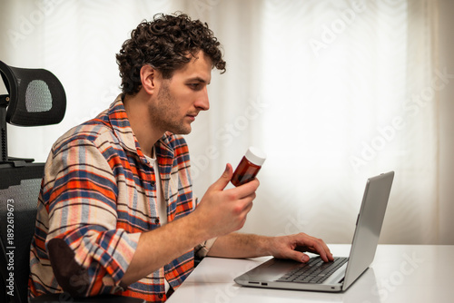 Wallpaper Mural Young man holding pill bottle and analyzing medical information on Internet via laptop while sitting comfortably in his living room at home.	 Torontodigital.ca