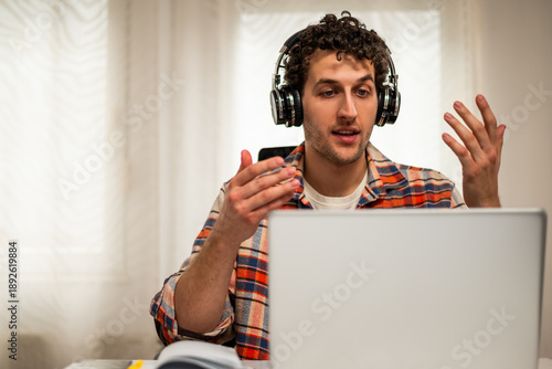 Wallpaper Mural Young man with wireless headphones talking and gesturing during a video call on laptop while sitting in his cozy home.	 Torontodigital.ca