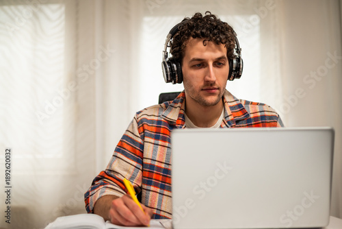 Wallpaper Mural Focused young man wearing wireless headphones, working on his laptop at home while taking notes on a notebook.	 Torontodigital.ca