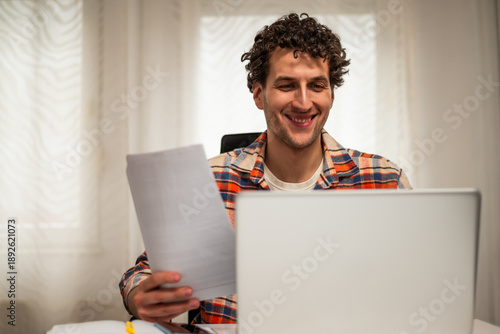 Wallpaper Mural Cheerful young freelancer using laptop and checking documents while working at his cozy home.	 Torontodigital.ca