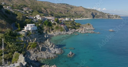 High-angle aerial view of a rugged coastline where colorful houses are built into the rocky cliffs, overlooking clear turquoise waters and a secluded beach nestled at the foot of a verdant hillside.