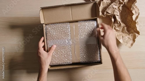 Eco friendly packaging scene shows hands using kraft paper cushioning as void fill around bubble wrapped product, preparing parcel for delivery with reduced plastic waste concept.