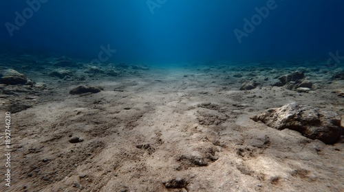 Serene underwater view of a clear blue ocean showing a sandy seabed with scattered rocks and gentle light filtering from above