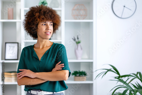 young elegant African American ethnic female with Afro curly hairstyle standing against blurred home or office interior background. crossed hands looking at right side, copy space