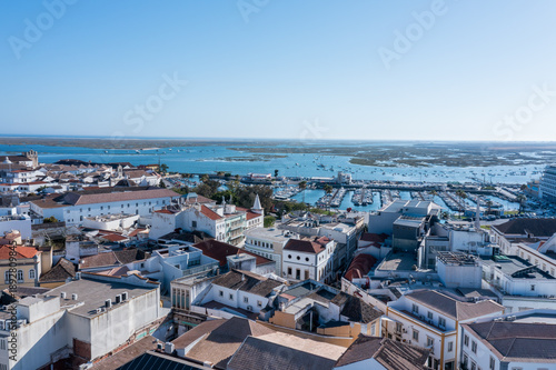 Tourists explore Faro's Old Town while boats float in the blue waters of Ria Formosa. The clear sky highlights the beauty of the Algarve region