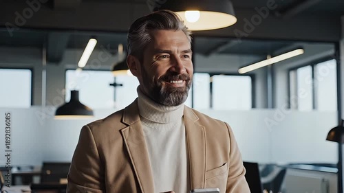 A stylish bearded businessman looking thoughtfully away while holding a smartphone in a sophisticated modern office.