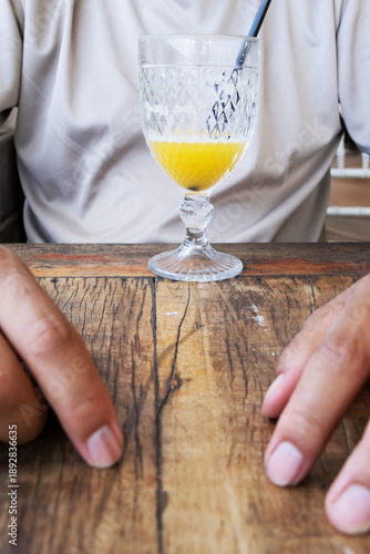 Image in portrait orientation showing part of an adult's hands on a wooden table, and at the end of the table, near the person's body, a glass with the remaining orange juice