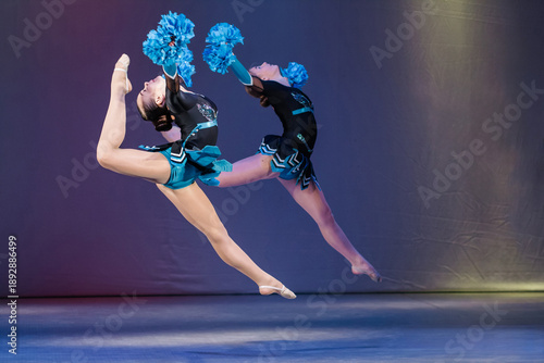 Two young cheerleaders leap gracefully in mid-air during a synchronized routine. Wearing black and turquoise uniforms with blue pom-poms, they perform a powerful jump on a lit stage