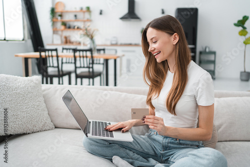 Young woman sitting relaxing on sofa in living room use laptop make online payment with credit card, female rest on couch at home shopping on internet, secure banking service system concept