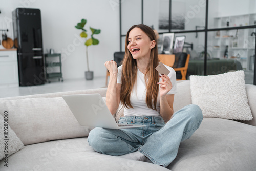 Emotional young Caucasian woman sitting on couch in living room and using laptop and credit card, checking new website for online shopping or banking, home interior, copy space, catching sales