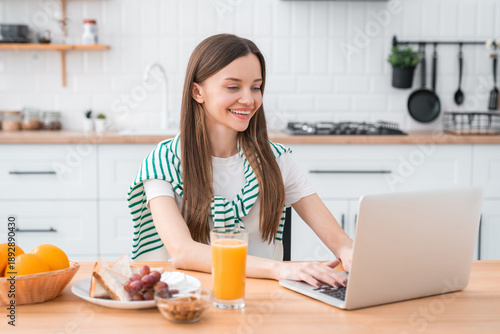Young woman working remotely at home kitchen table with laptop healthy breakfast, representing freelance lifestyle, flexible schedule, and modern work-from-home concept.
