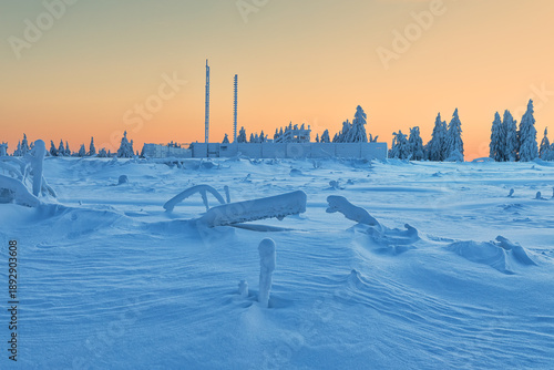 A frozen plateau with sculpted snow drifts and icy forms surrounds a distant facility beneath a glowing pastel sky.