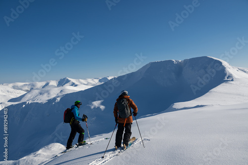 Fotografie Two skiers traverse a snowy alpine ridge in clear blue sky during an off-piste a