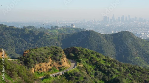 Scenic view of the Griffith Observatory and downtown Los Angeles skyline from a popular hiking trail in the Hollywood Hills on a sunny, hazy day.
