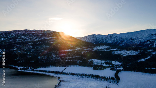 Aerial view of icy waters reflect the muted sky, embraced by snow-laden mountains and frosted forests, a landscape of serene contrast In Norway. Toma Aerea incredible de paisaje nevado.
