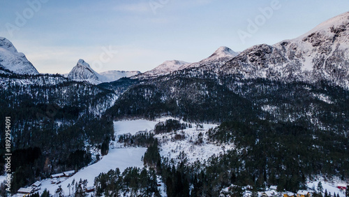 Aerial view of Reine under a blanket of fresh snow, nestled beneath the majestic, cloud-kissed mountains, a serene winter tableau of dark waters and bright rooftops, Reine, Nordland, Stryn, Norway.